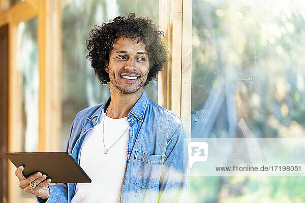 Lächelnder Mann mit digitalem Tablet  der wegschaut  während er sich an ein Fenster im Vorgarten lehnt