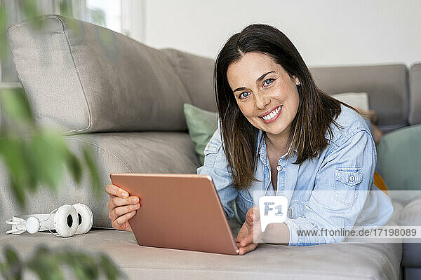 Schöne Frau lächelt  während sie ein digitales Tablet auf dem Sofa zu Hause hält