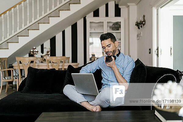 Geschäftsmann mit Laptop  der zu Hause auf dem Sofa sitzt und mit seinem Handy telefoniert