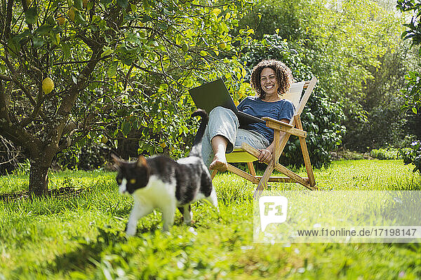 Glückliche Frau mit Laptop  die ihre Katze betrachtet  während sie auf einem Liegestuhl am Zitronenbaum sitzt