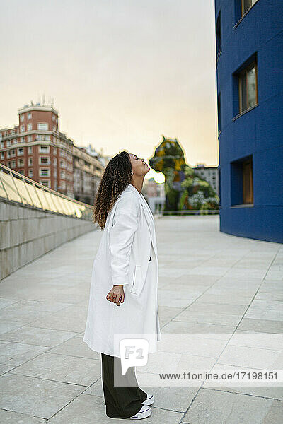 Young woman wearing overcoat while standing on footpath during sunset