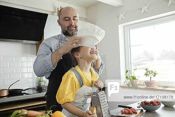 Father putting chef's hat on daughter head while standing in kitchen at home