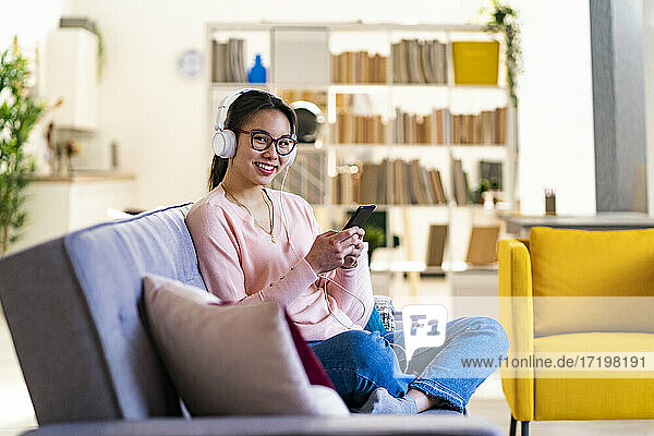 Junge Frau mit Mobiltelefon und Kopfhörern sitzt zu Hause auf dem Sofa