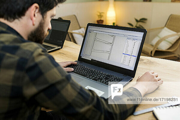 Männlicher Schreiner  der am Schreibtisch sitzend eine Zeichnung auf einem Laptop anfertigt