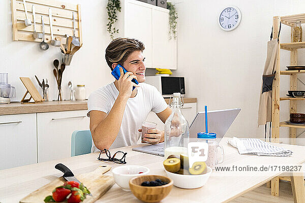Lächelnder junger Mann  der mit dem Handy telefoniert  während er mit dem Laptop in der Küche zu Hause sitzt