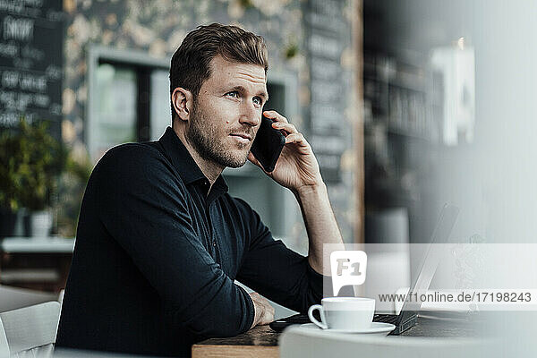 Businessman talking on smart phone while sitting by laptop at cafe