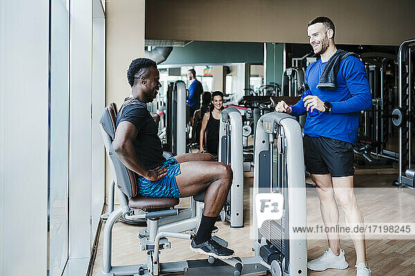 Lächelnde männliche Freunde unterhalten sich an einem Fitnessgerät im Fitnessstudio