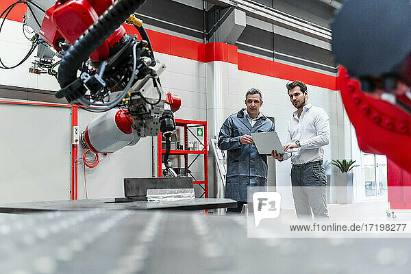 Männliche Ingenieure mit Laptop  die in einer Fabrik stehen und sich mit Robotik beschäftigen