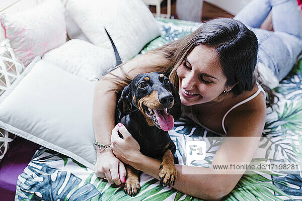 Smiling woman embracing dog while lying on front on bed at home