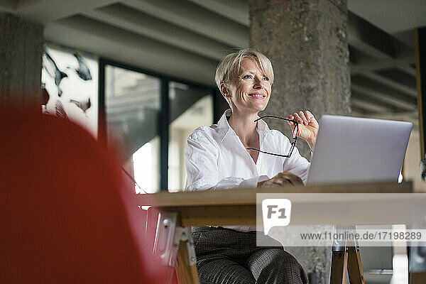 Lächelnde Geschäftsfrau mit Laptop  die eine Brille hält  während sie am Schreibtisch im Heimbüro sitzt
