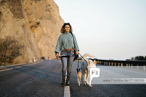 Smiling woman with Siberian Husky and Jack Russell Terrier looking away while walking on mountain road against sky