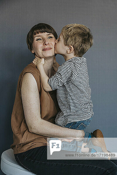 Cute boy kissing mother on cheek over gray background