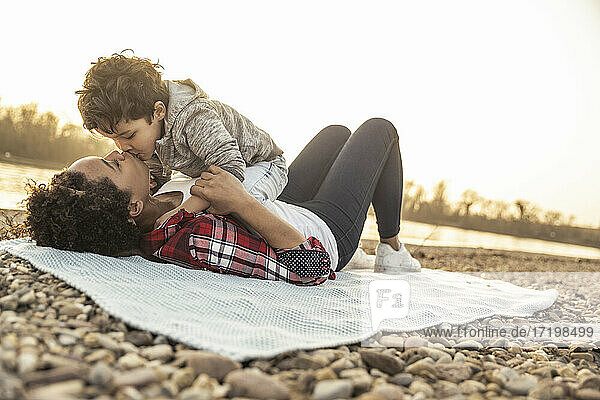 Junge küsst Mutter auf Decke über Kieselsteinen bei Sonnenuntergang