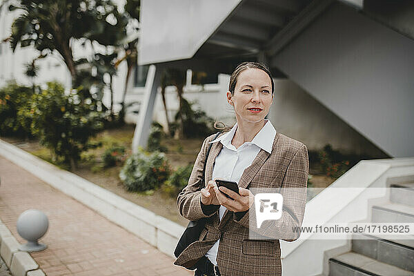 Geschäftsfrau hält Smartphone und schaut weg  während sie vor einem Bürogebäude steht