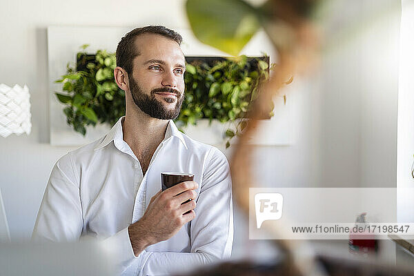 Nachdenklicher Unternehmer  der lächelnd eine Kaffeetasse im Büro hält
