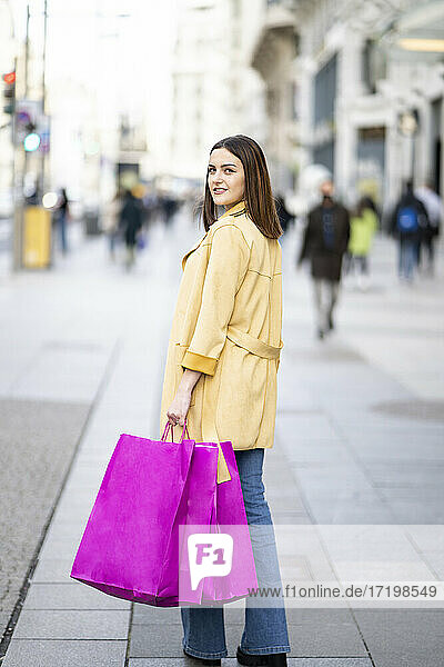 Junge Frau mit Einkaufstüten  die über die Schulter schaut  während sie auf einem Fußweg in der Stadt spazieren geht
