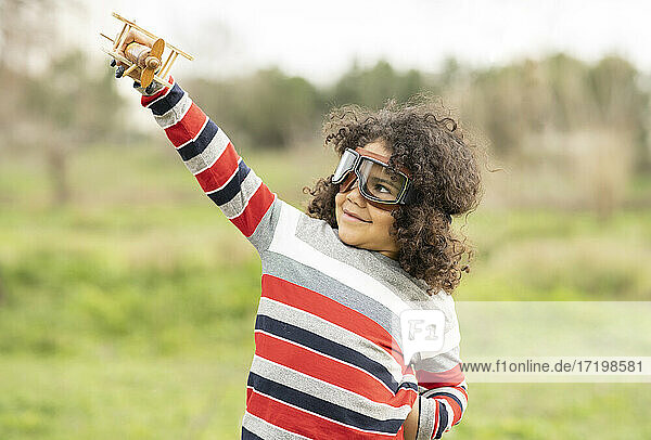 Niedlicher Junge mit Fliegerbrille  der mit einem Holzspielzeugflugzeug spielt