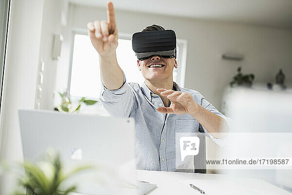 Lächelnder männlicher Fachmann  der einen Virtual-Reality-Simulator trägt und gestikuliert  während er in seinem Büro sitzt