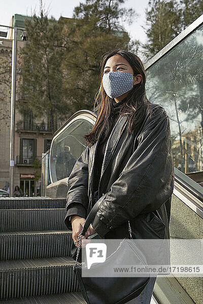 Asiatische Frau mit Handtasche  die während der Pandemie auf einer Rolltreppe steht und wegschaut
