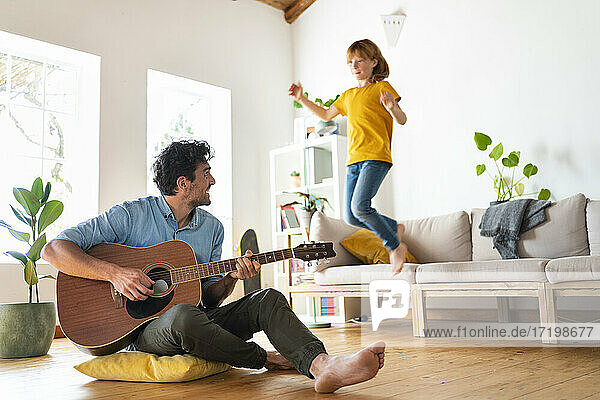 Father playing guitar while daughter jumping from sofa in living room at home