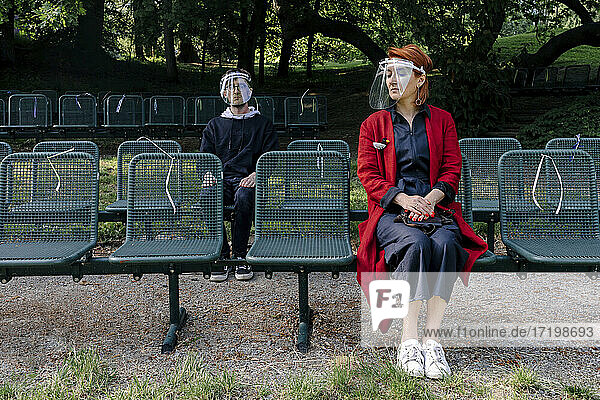 Mutter und Sohn mit Gesichtsschutz auf einem Sitz im Park während COVID-19