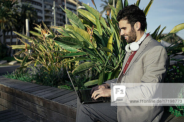 Smiling businessman using laptop while sitting on retaining wall