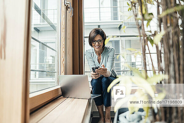 Glückliche Geschäftsfrau  die im Büro sitzend eine Textnachricht über ihr Smartphone verschickt