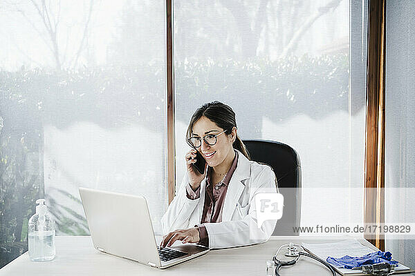 Eine Ärztin benutzt einen Laptop  während sie im Krankenhaus mit einem Mobiltelefon spricht