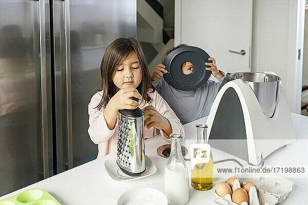 Nettes Mädchen  das eine Reibe hält  während ihr Bruder in der Küche mit einem Utensil spielt