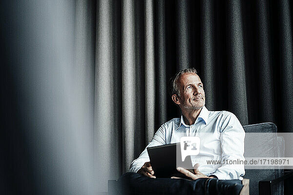 Nachdenklicher Geschäftsmann mit digitalem Tablet  der wegschaut  während er auf einem Sessel in einer Büro-Cafeteria sitzt