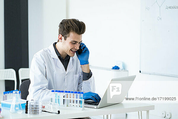 Männlicher Wissenschaftler  der mit einem Smartphone spricht  während er einen Laptop am Schreibtisch im Labor benutzt