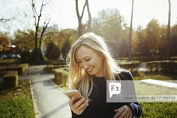 Blonde Frau lacht  während sie ihr Smartphone im Park verklagt