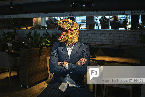 Businessman wearing dinosaur mask while sitting with arms crossed in cafe