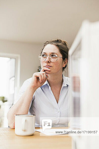 Thoughtful female entrepreneur wearing eyeglasses sitting at desk looking away