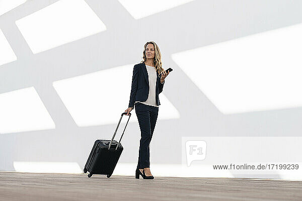 Businesswoman looking away while walking with mobile phone and luggage against white wall