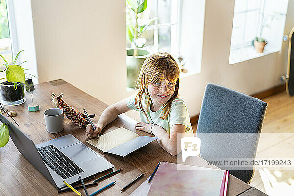 Smiling redhead girl looking away while doing homework in front of laptop at home