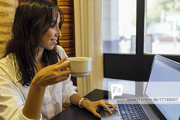 Junge Geschäftsfrau arbeitet am Laptop und hält eine Tasse Kaffee in einem Café