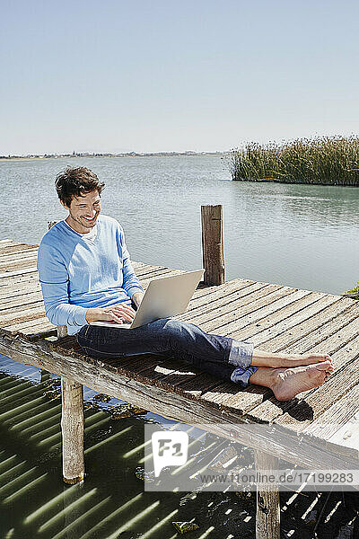Lächelnder Mann  der einen Laptop benutzt  während er an einem sonnigen Tag auf einem Steg sitzt