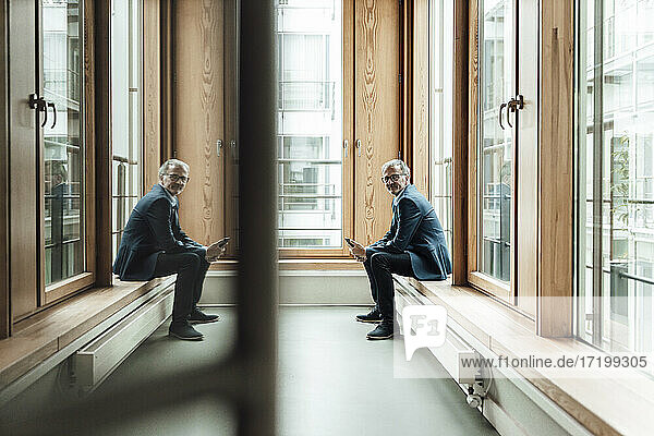 Businessman with mobile phone sitting at glass window in office corridor