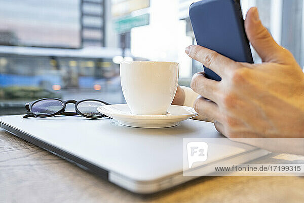 Die Hand eines Geschäftsmannes  der ein Smartphone benutzt  während er einen Kaffee mit einem Laptop am Kaffeetisch trinkt