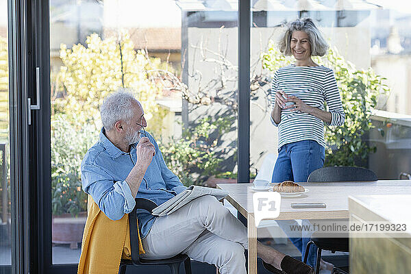 Älterer Mann mit Zeitung am Tisch sitzend mit Frau im Hintergrund zu Hause stehend