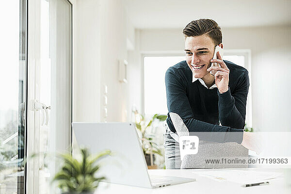 Male entrepreneur talking on phone while leaning on chair at home office