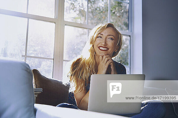 Lächelnde Frau mit Laptop  die wegschaut  während sie zu Hause sitzt