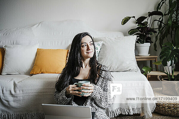 Lächelnde Frau mit Laptop  die eine Kaffeetasse hält und auf dem Boden vor dem Sofa im Wohnzimmer sitzt