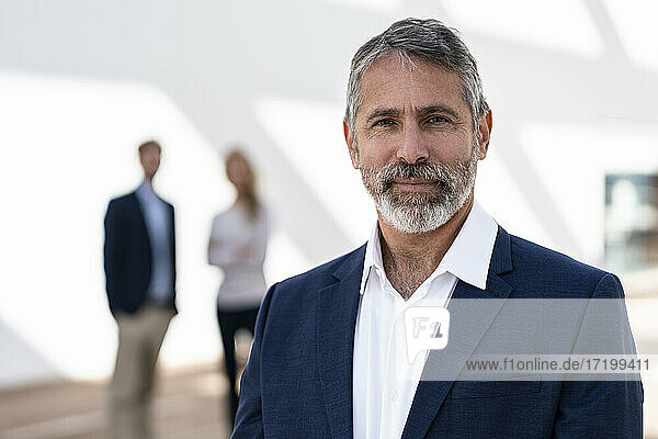 Mature businessman standing with colleagues in background at office terrace