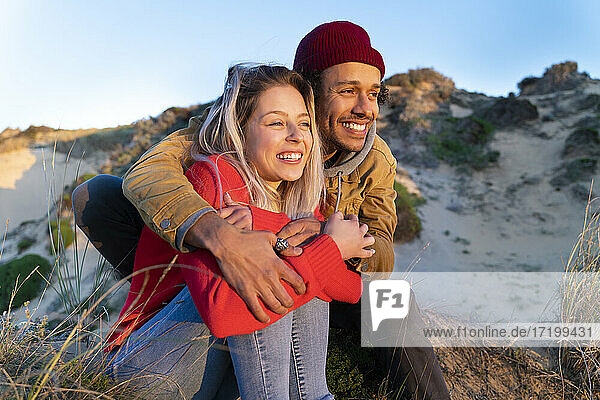 Mann mit Strickmütze  der eine schöne Frau umarmt  während er auf einer Sanddüne sitzt