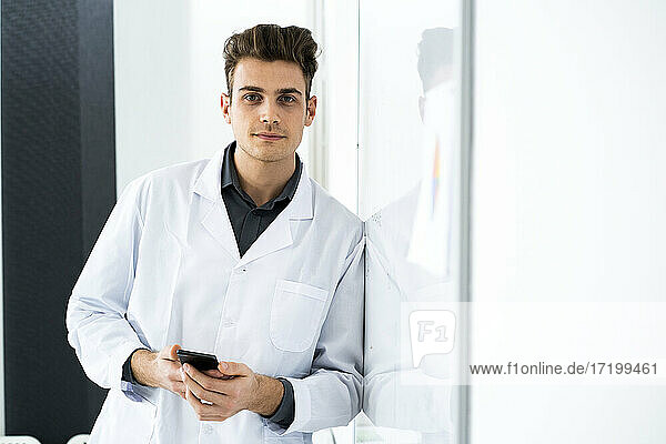 Handsome scientist with smart phone leaning on whiteboard in laboratory