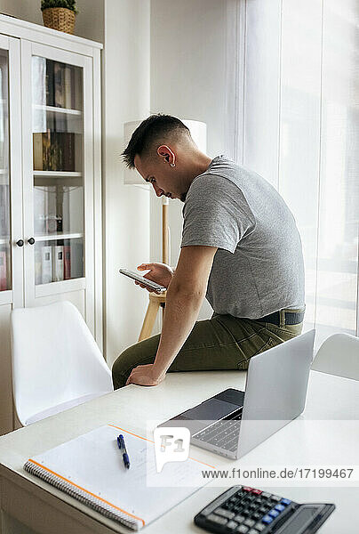 Männlicher Unternehmer  der ein Smartphone benutzt  während er im Büro zu Hause am Schreibtisch neben dem Laptop sitzt