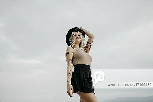 Tattooed young woman wearing hat standing against sky