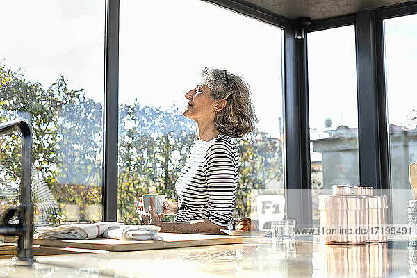 Sonnenlicht fällt auf eine ältere Frau  die mit einer Kaffeetasse in der Küche steht